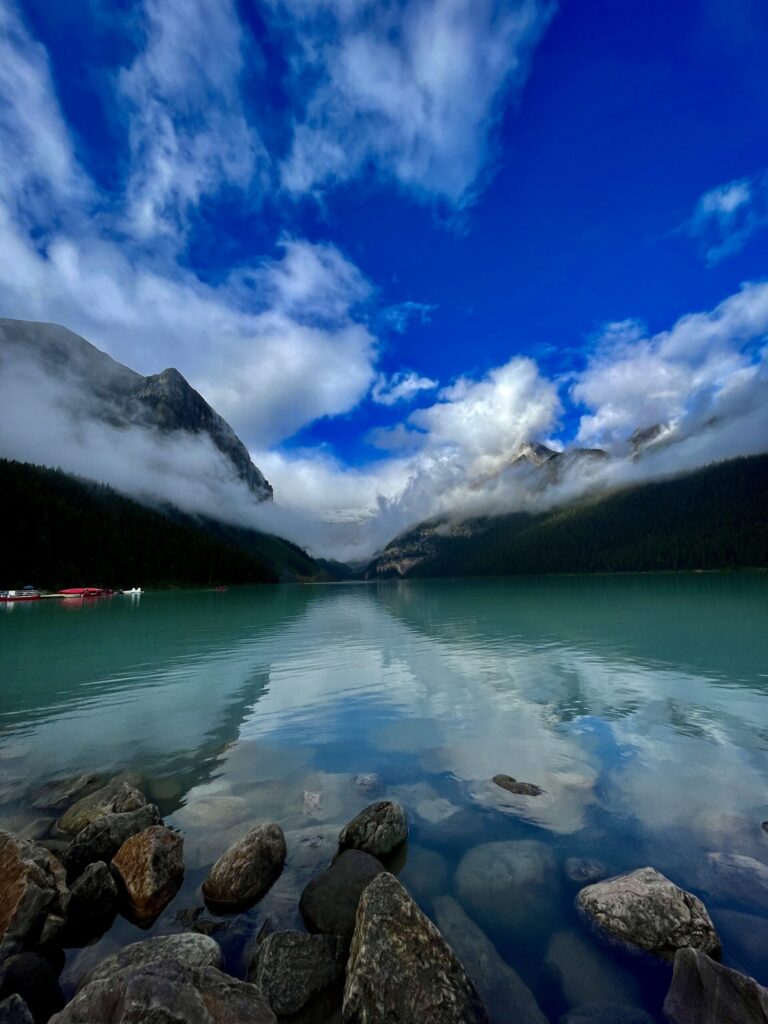 Turquoise alpine lake surrounded by forested mountains with mist-covered peaks under a blue sky, red canoes moored on the left shore
