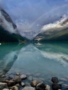 Turquoise alpine lake surrounded by forested mountains with misty peaks and a rainbow arching across the sky, rocks visible in the foreground water