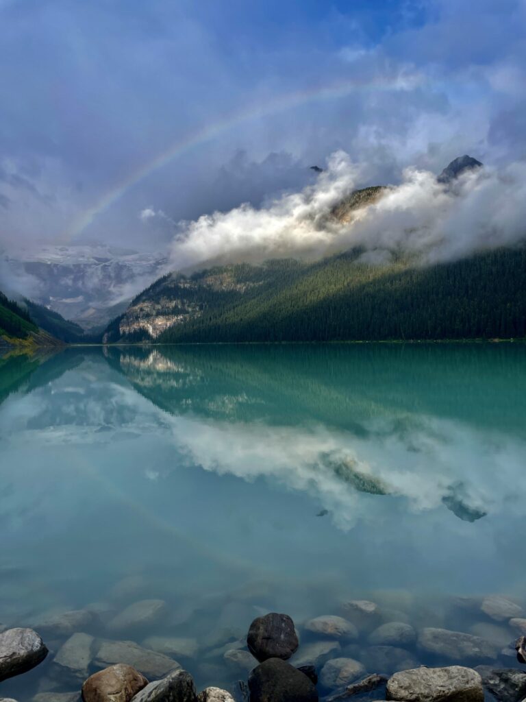 Turquoise alpine lake with forested mountains shrouded in mist and low clouds, rocky shoreline in foreground, distant valley visible through clearing weather