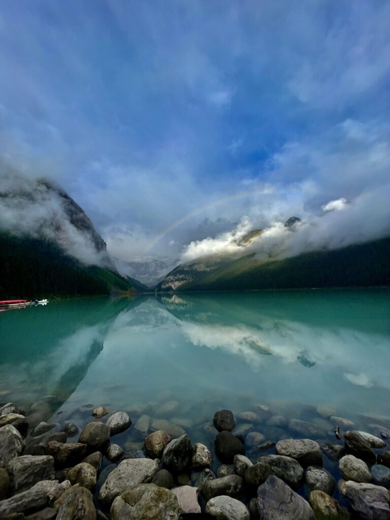 Turquoise alpine lake with forested mountains shrouded in mist, rocky shoreline in foreground, and red canoe visible on left shore