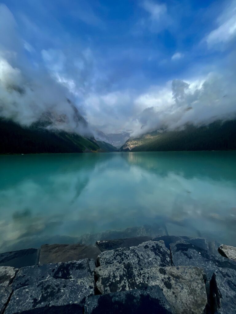 Turquoise alpine lake surrounded by forested mountains and mist, with rocky shoreline in foreground and cloudy sky above