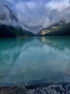 Turquoise alpine lake surrounded by forested mountains shrouded in mist and low clouds, with snow-capped peaks visible in the distance