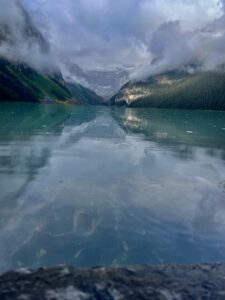 Turquoise alpine lake surrounded by forested mountains with low-hanging clouds and mist, reflecting the sky and peaks in still water