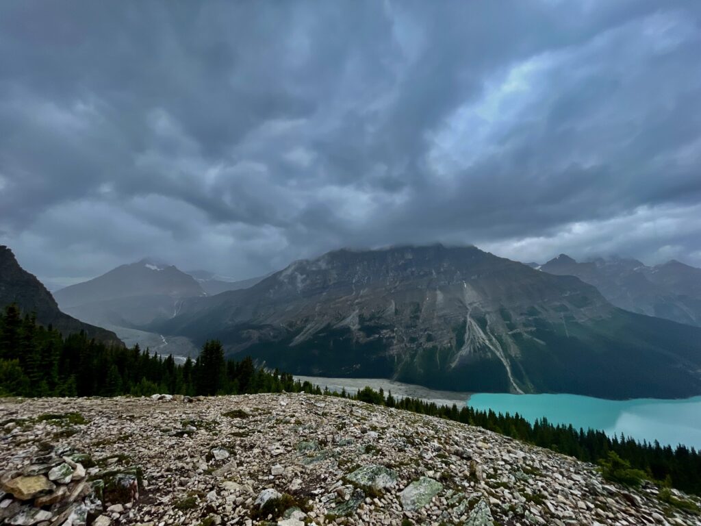 Turquoise glacial lake surrounded by steep rocky mountains, coniferous forest, and dramatic cloudy sky in an alpine landscape