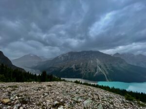 Turquoise glacial lake surrounded by steep rocky mountains, coniferous forest, and dramatic cloudy sky in an alpine landscape