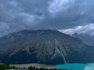 Dramatic mountain landscape with steep rocky peaks, forested slopes, and turquoise glacial lake under stormy sky with dark clouds