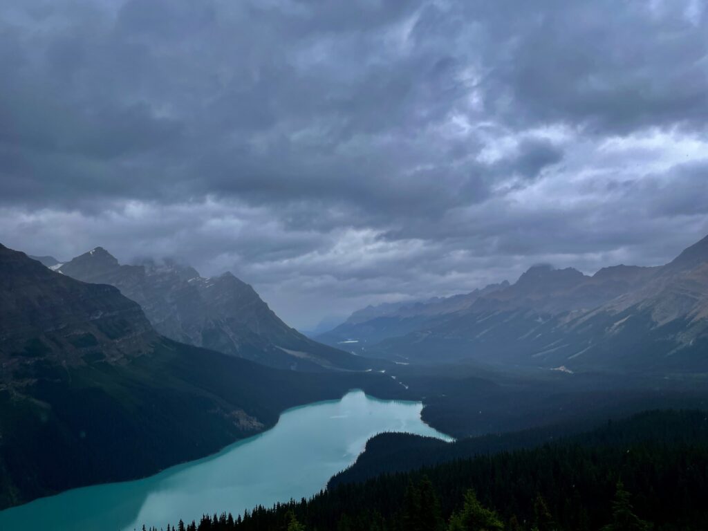 Turquoise glacial lake nestled in a mountain valley under dramatic cloudy skies, surrounded by forested peaks and snow-capped ridges