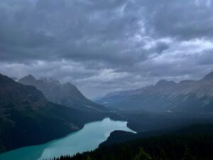 Turquoise glacial lake nestled in a mountain valley under dramatic cloudy skies, surrounded by forested peaks and snow-capped ridges