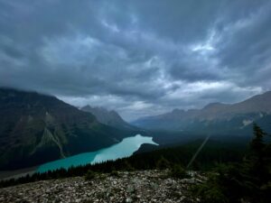 Turquoise glacial lake nestled in a mountain valley under dramatic cloudy skies, surrounded by forested peaks and rocky terrain