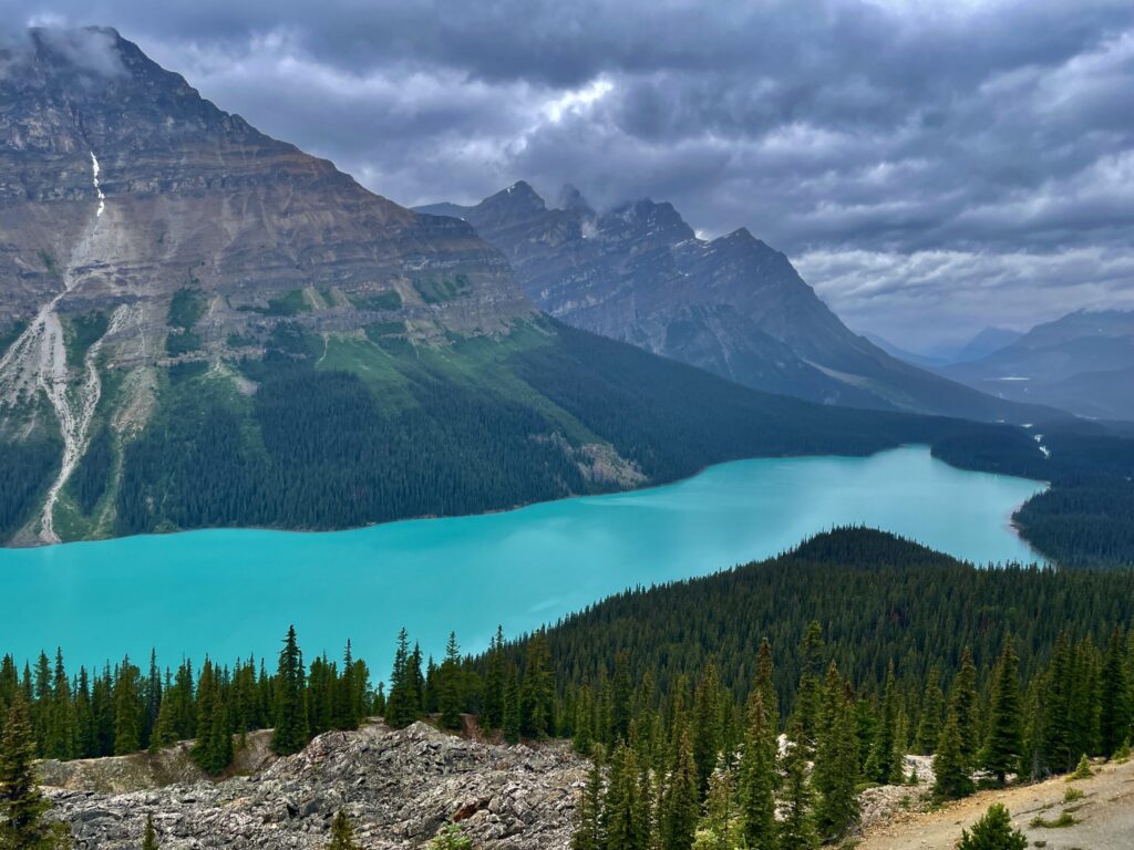 Turquoise alpine lake surrounded by coniferous forest, rocky peaks, and a waterfall cascading down the mountainside under a cloudy sky