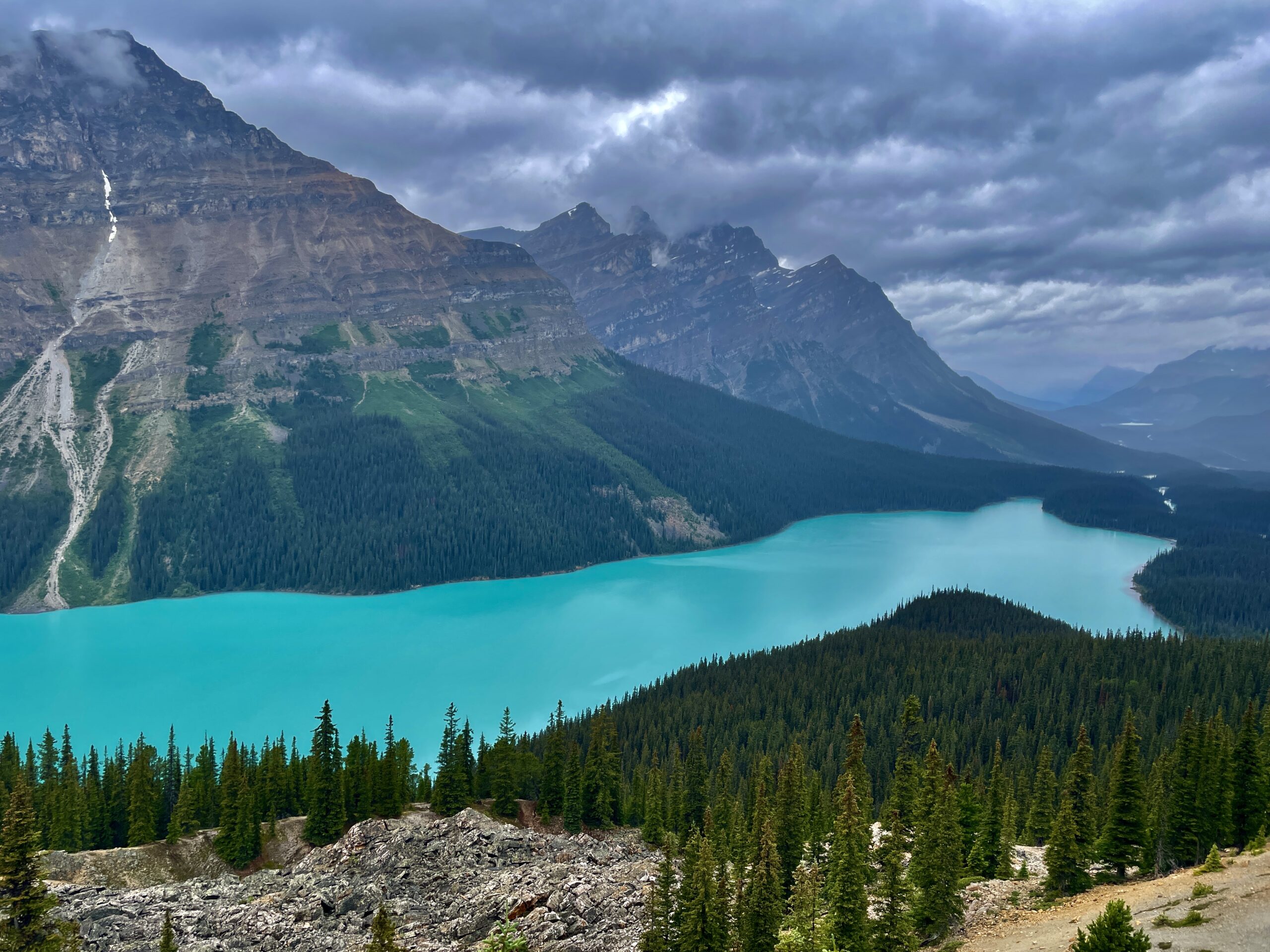 Turquoise alpine lake surrounded by coniferous forest, rocky peaks, and a waterfall cascading down the mountainside under a cloudy sky