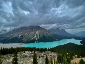 Turquoise alpine lake nestled in a mountain valley with rocky peaks, coniferous forest, and dramatic cloudy sky overhead