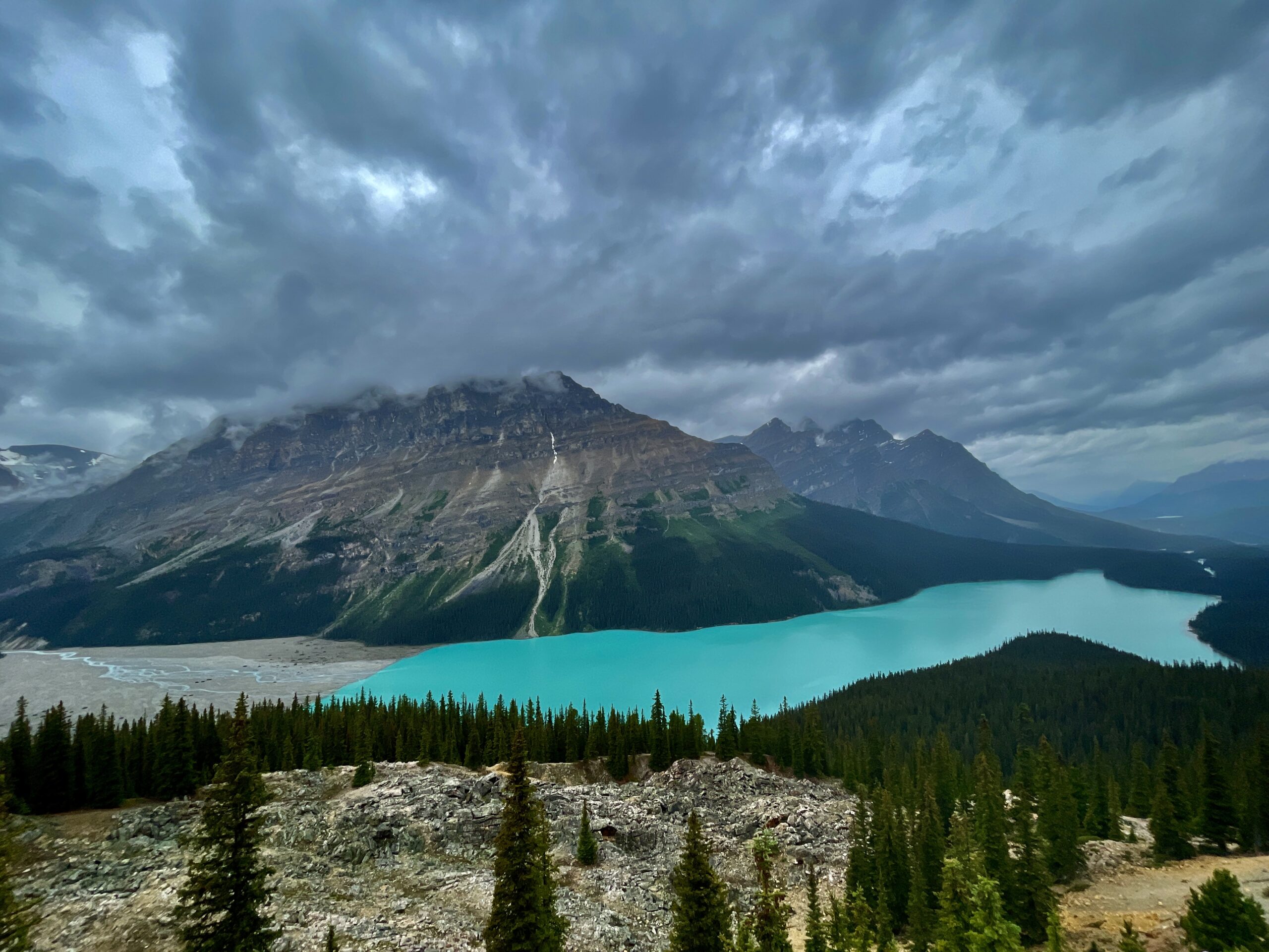 Turquoise alpine lake nestled in a mountain valley with rocky peaks, coniferous forest, and dramatic cloudy sky overhead