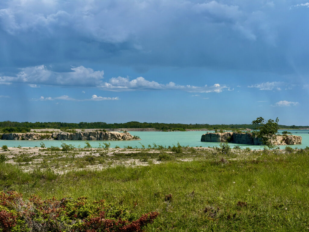Turquoise quarry lake surrounded by limestone cliffs, forested shoreline, and grassy meadow under blue sky with white clouds