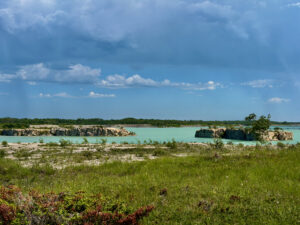 Turquoise quarry lake surrounded by limestone cliffs, forested shoreline, and grassy meadow under blue sky with white clouds