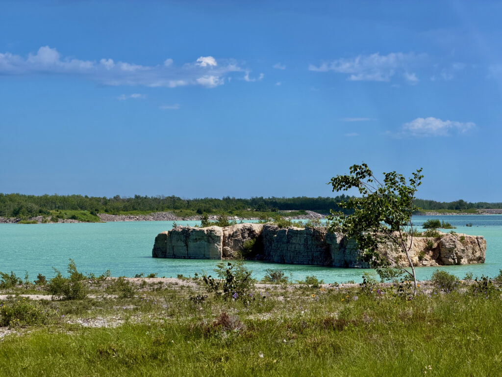 Turquoise water with rocky outcroppings and forested shoreline under clear blue sky with white clouds