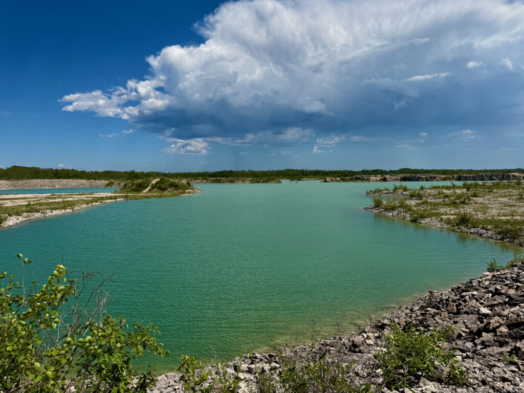 Turquoise quarry lake surrounded by rocky limestone shores, forested banks, and white clouds in blue sky