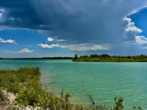 Turquoise lagoon with mangrove vegetation along the shoreline, forested islands in the distance, and blue sky with white and grey clouds