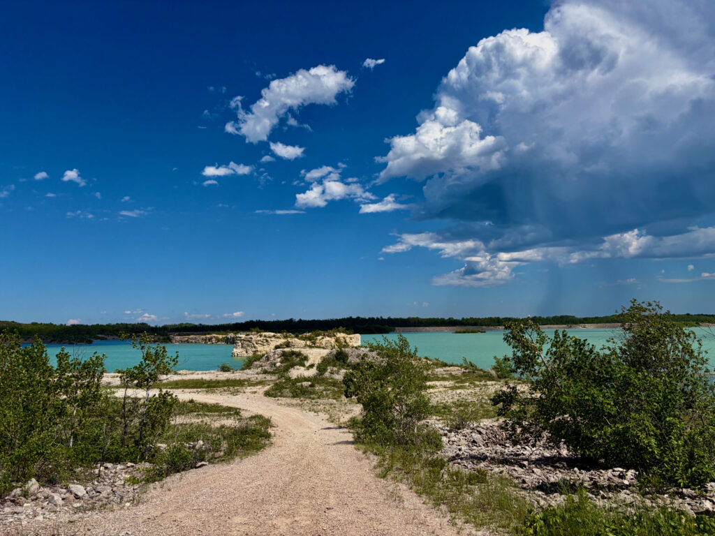 Turquoise quarry lake surrounded by sandy terrain, limestone cliffs, and vegetation under a blue sky with white clouds