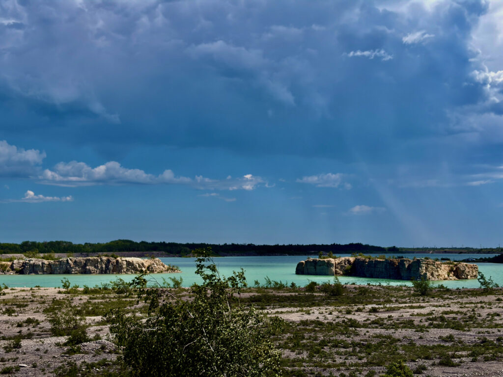 Turquoise quarry lake surrounded by limestone cliffs and forested shoreline under a blue sky with white clouds
