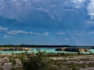 Turquoise quarry lake surrounded by limestone cliffs and forested shoreline under a blue sky with white clouds