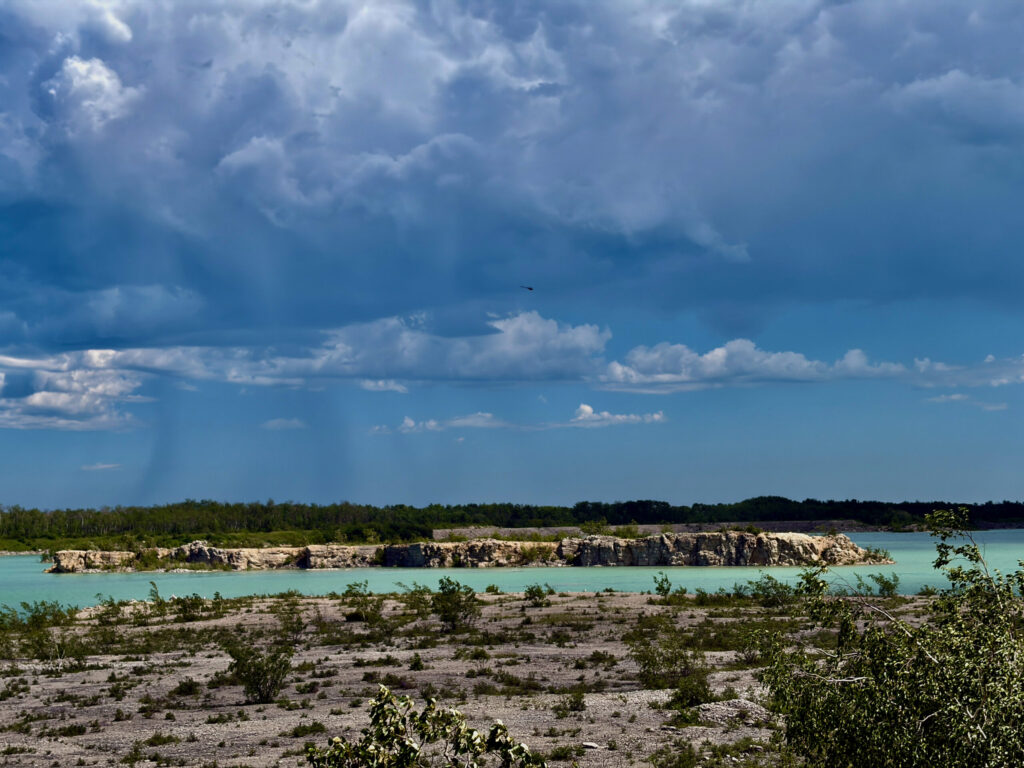 Turquoise quarry lake with limestone cliffs, forested shoreline, and dramatic cloudy sky with crepuscular rays