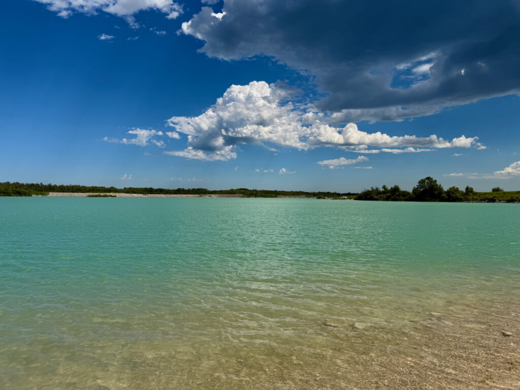 Turquoise lake with sandy beach, forested shoreline, and blue sky with white clouds