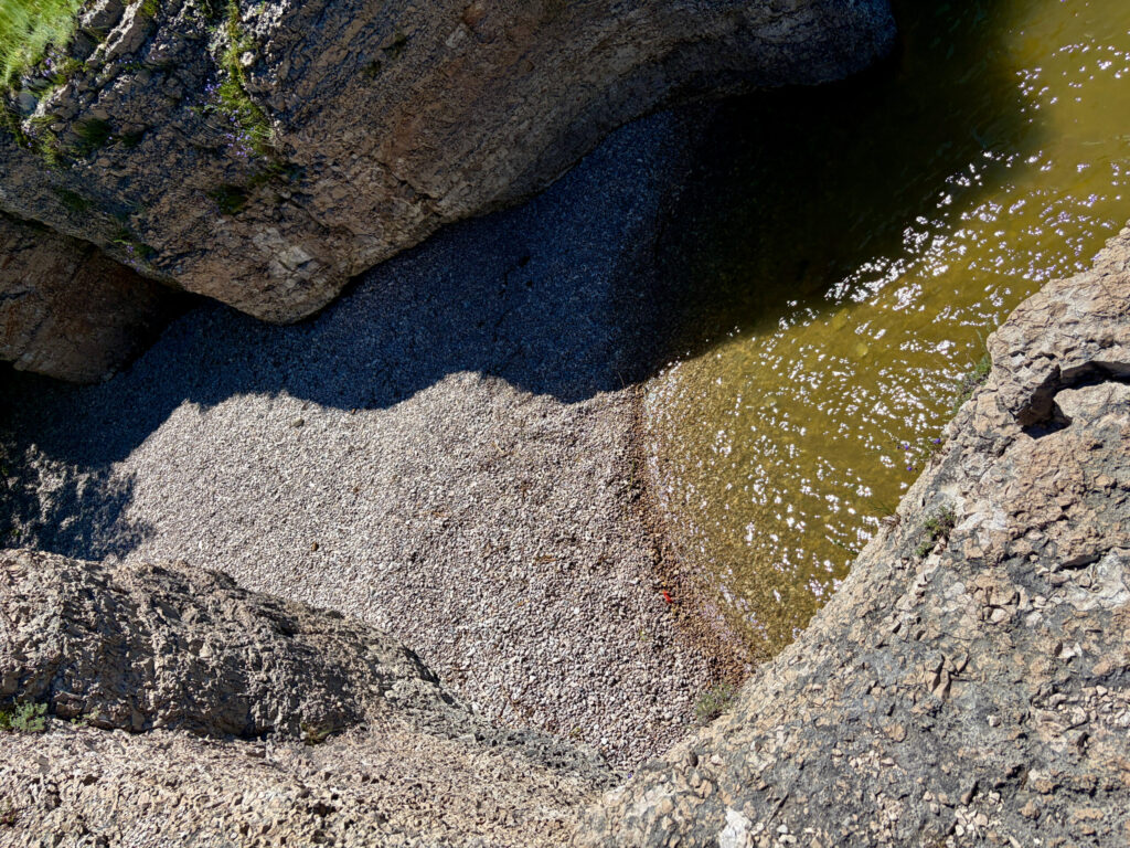 Aerial view of a rocky tidal pool with dark blue water, pale pebbled beach, and moss-covered stone formations along the shoreline