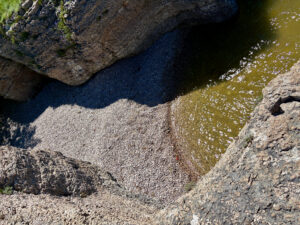 Aerial view of a rocky tidal pool with dark blue water, pale pebbled beach, and moss-covered stone formations along the shoreline