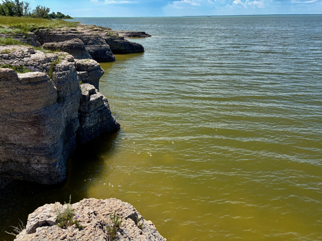 Rocky shoreline with layered limestone cliffs overlooking calm greenish-brown water under blue sky with white clouds