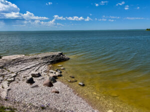 Rocky shoreline with shallow greenish-yellow water extending to a calm horizon under blue sky with white clouds