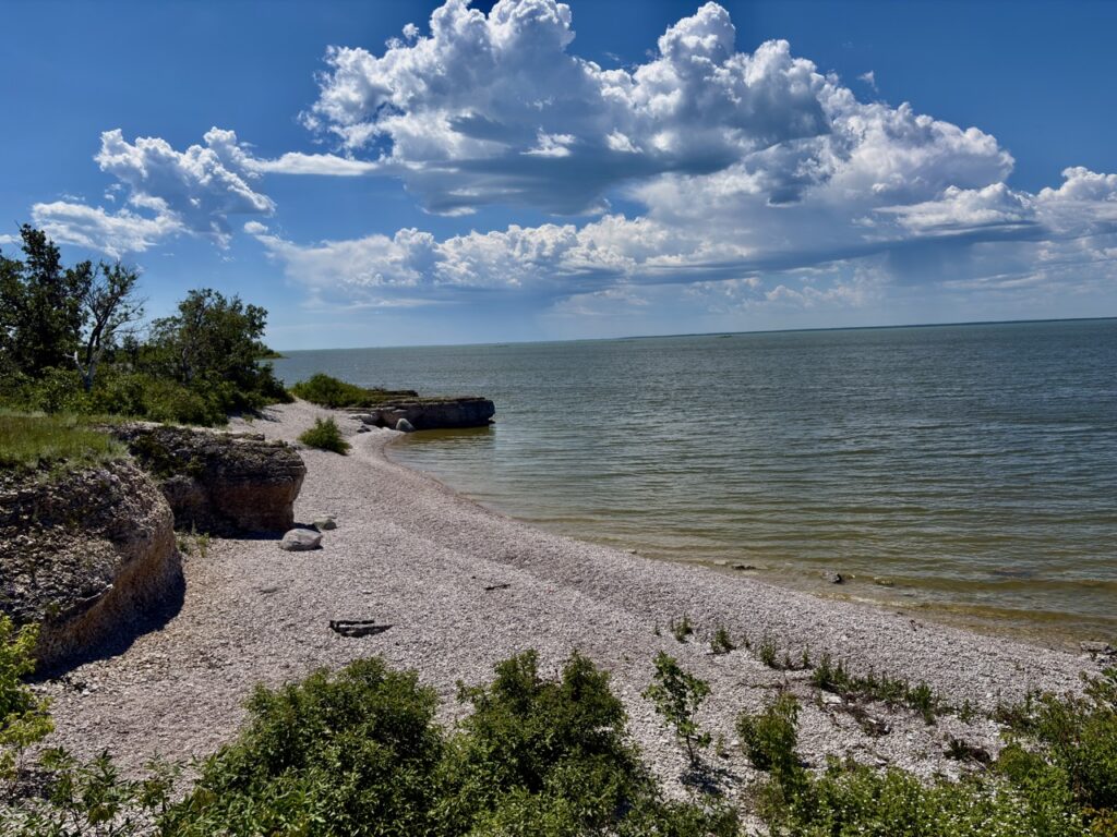 Rocky shoreline with pebble beach, green vegetation, and calm water under a blue sky with white clouds