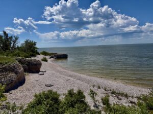 Rocky shoreline with pebble beach, green vegetation, and calm water under a blue sky with white clouds