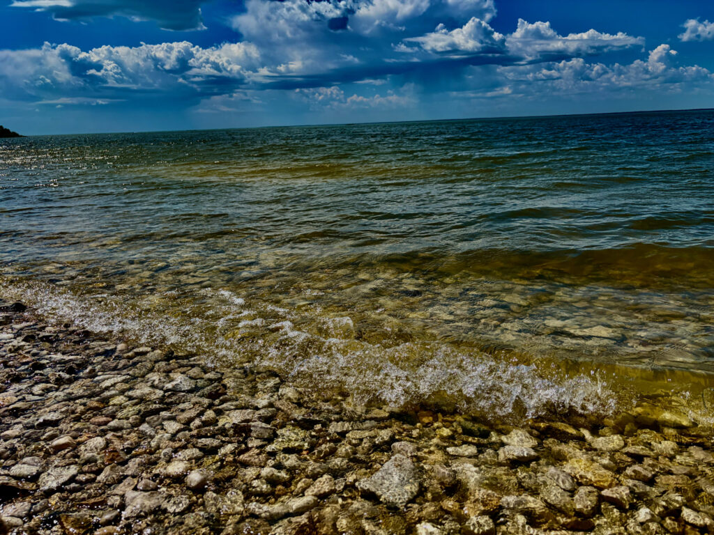 Clear shallow water with rocky shoreline and waves under a blue sky with white clouds
