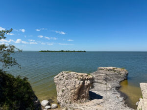 Rocky shoreline with limestone formations in shallow turquoise water, forested island visible on the horizon under clear blue sky
