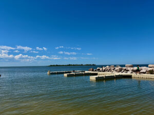 Concrete dock extending into calm blue water with rocky breakwater, forested island in distance, and clear sky with white clouds
