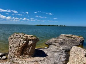 Rocky shoreline with limestone formations in shallow turquoise water, distant forested island, and clear blue sky with white clouds
