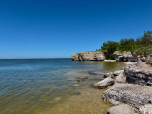 Rocky shoreline with limestone formations and clear blue water under a cloudless sky, a person visible on the beach in the distance
