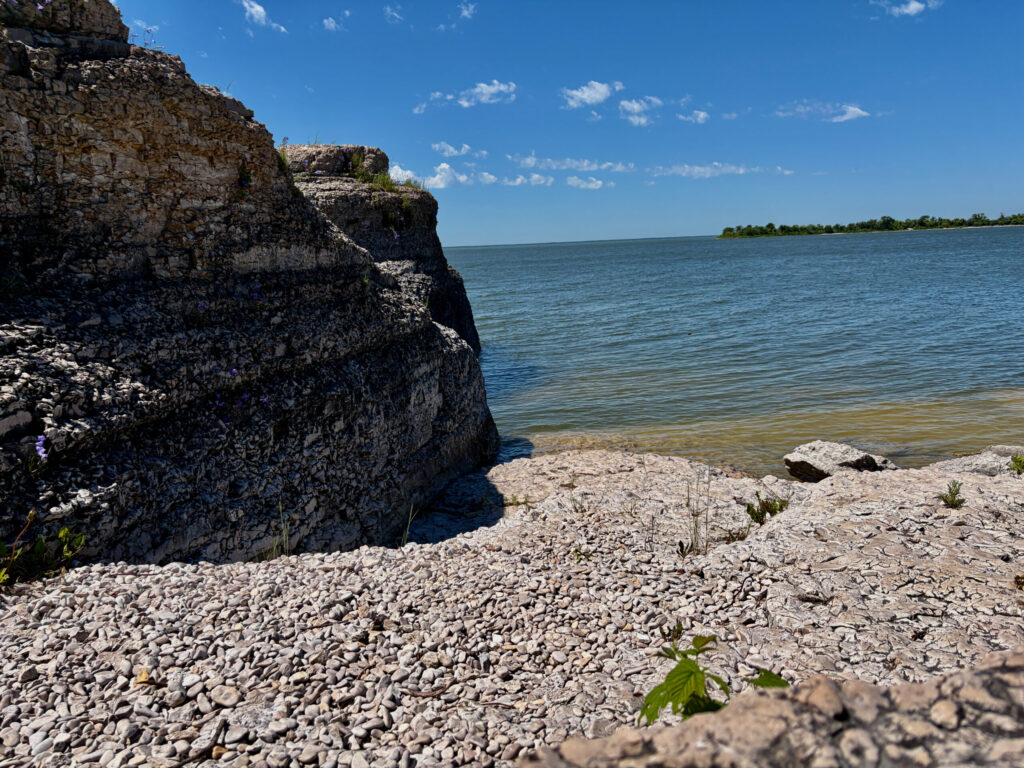 Rocky shoreline with limestone cliffs overlooking calm blue-green water, sandy beach, and distant tree-lined shore under clear sky