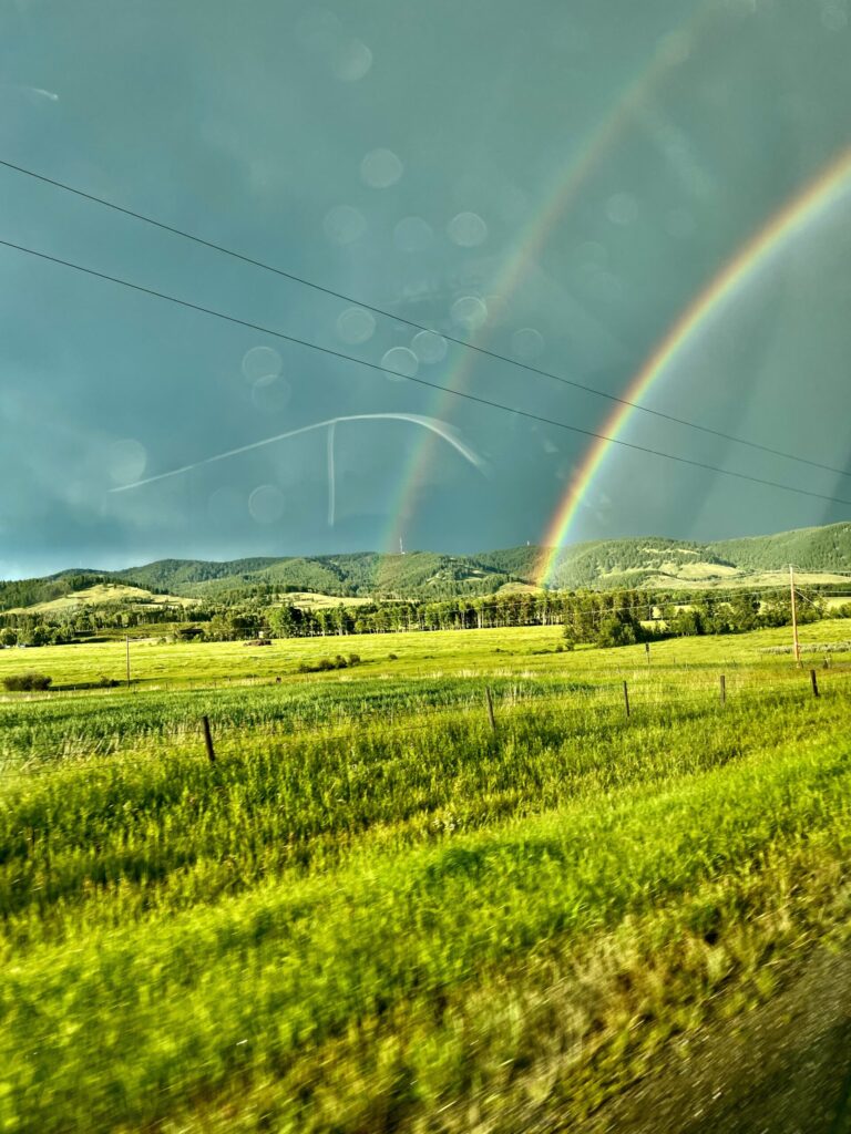 Double rainbow over a lush green valley with forested hills, pasture fencing, and power lines under a clearing sky