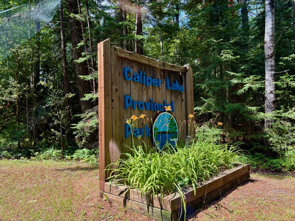 Caliper Lake Provincial Park sign with blue text and lake illustration on wooden board surrounded by forest