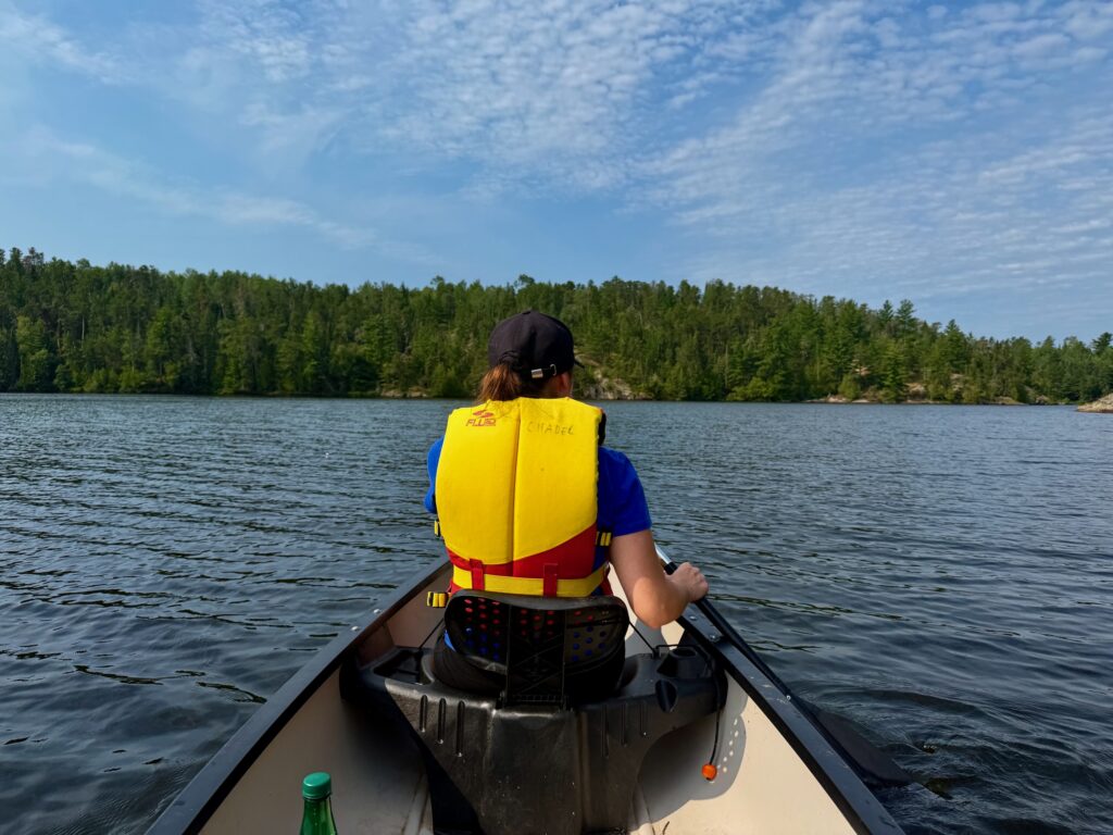 Person wearing a yellow life jacket paddling a canoe on a lake surrounded by coniferous forest under a blue sky