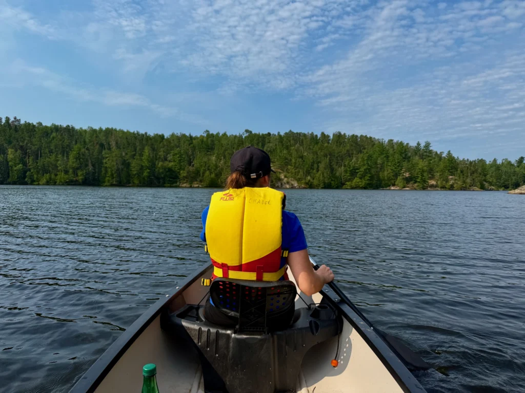Person wearing a yellow life jacket paddling a canoe on a lake surrounded by coniferous forest under a blue sky