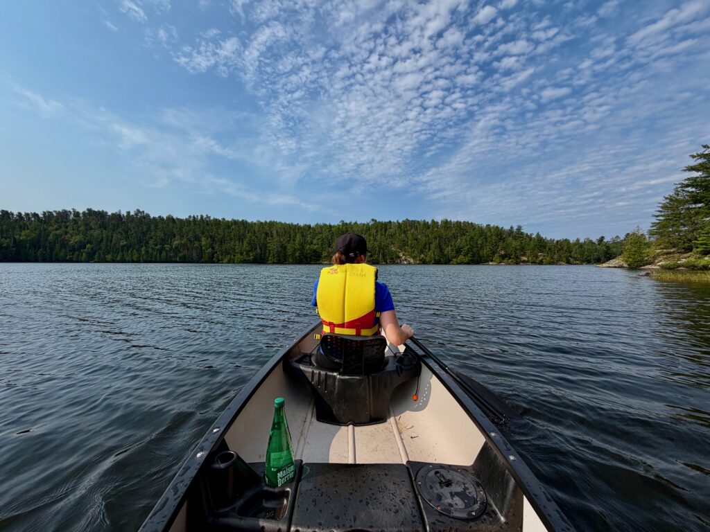 Person in yellow life jacket canoeing on a calm lake surrounded by evergreen forest under a blue sky with wispy clouds