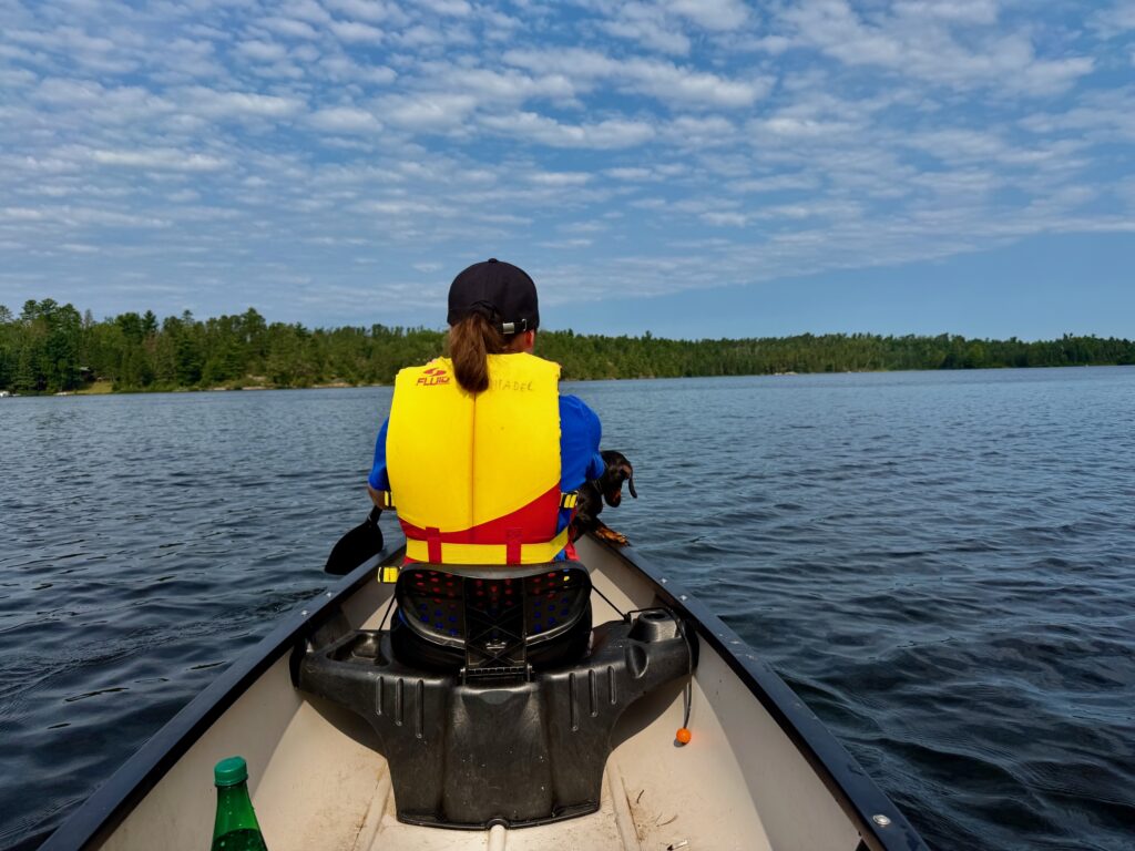 Person wearing a yellow and red life jacket and black cap sitting in the bow of a canoe on a calm lake, with a forested shoreline in the background