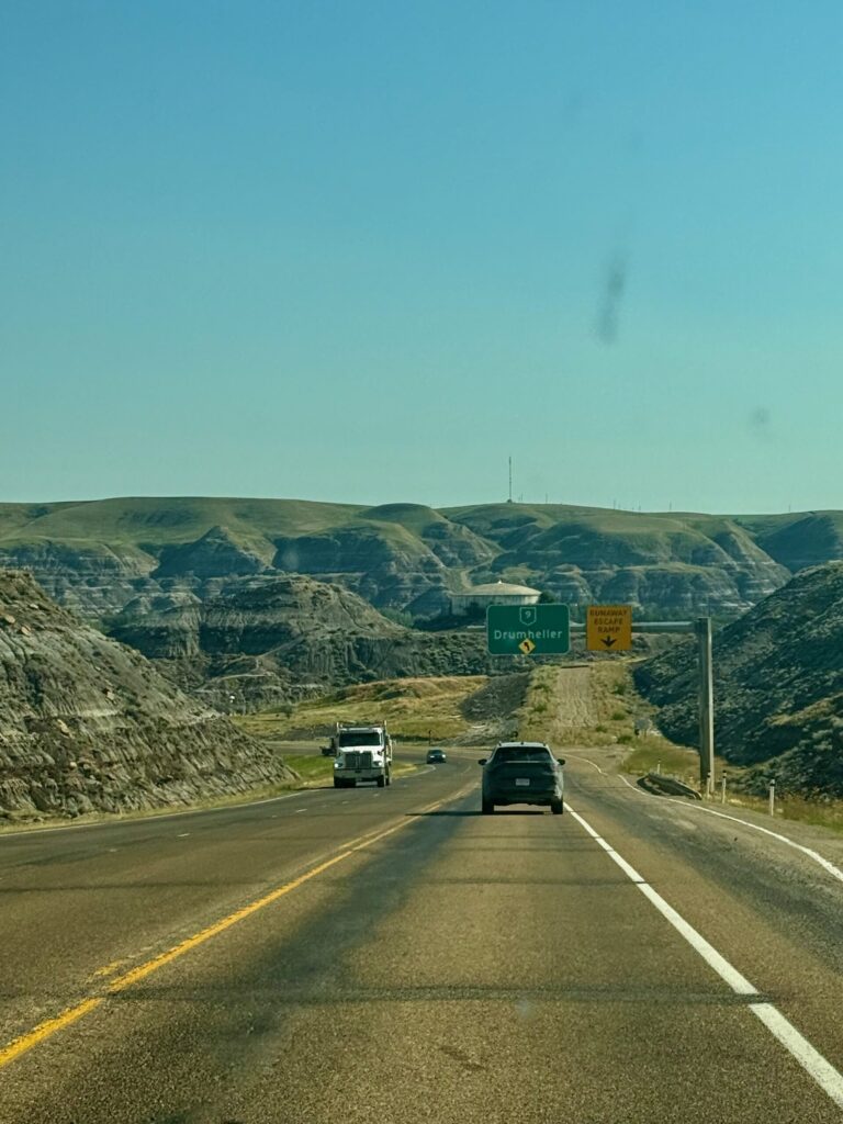 Highway approaching Drumheller with directional signs and deeply eroded badlands terrain on both sides of the road