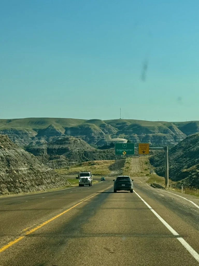 Highway approaching Drumheller with directional signs and deeply eroded badlands terrain on both sides of the road