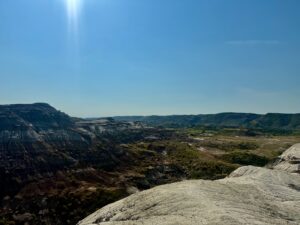 Expansive badlands landscape with layered rock formations, eroded valleys, scattered vegetation, and clear blue sky