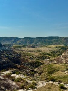 Panoramic view of a wide valley surrounded by layered badlands hills with exposed rock formations, sparse vegetation, and a winding river under clear blue sky