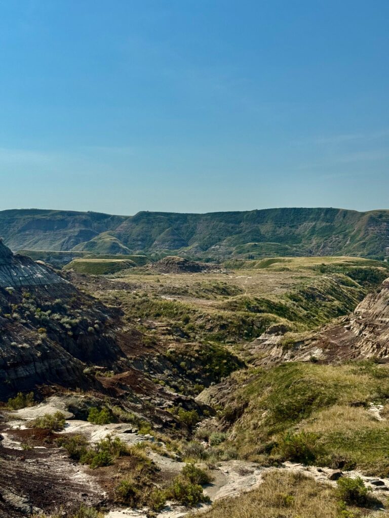 Panoramic view of a wide valley surrounded by layered badlands hills with exposed rock formations, sparse vegetation, and a winding river under clear blue sky